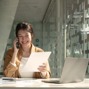 Charming Asian woman with a smile standing holding papers and mobile phone at the office.